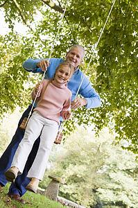 Grandfather pushing granddaughter on swing and smiling (selective focus)