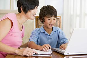 Woman helping young boy with laptop do homework in dining room smiling