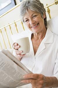 Woman in bedroom with coffee and newspaper smiling