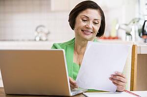 Woman in kitchen with laptop and paperwork smiling