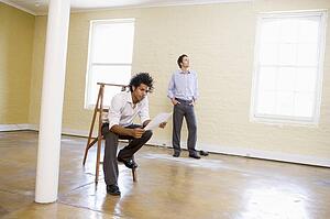 Man sitting on ladder in empty space holding paper with other man behind him