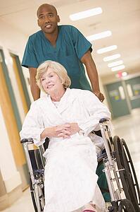 An Orderly Pushing A Senior Woman In A Wheelchair Down A Hospital Corridor