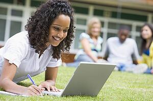 Student outdoors on lawn using laptop with other students in background (selective focus)