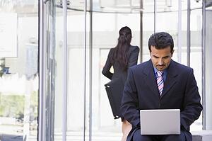 Businessman outdoors in front of building using laptop with businesswoman in background (high key/selective focus)