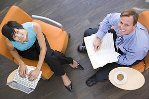 Two businesspeople sitting indoors with coffee laptop and folder smiling