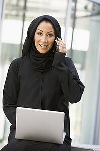 Businesswoman sitting outdoors by building with laptop using cellular phone smiling (selective focus)