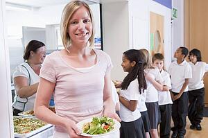 Students in cafeteria line with teacher holding her healthy meal and looking at camera (depth of field)