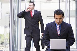 Businessman outdoors in front of building using laptop with businessman in background on cellular phone (high key/selective focus)