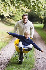 Man and young boy walking on path outdoors with wheelbarrow smiling (selective focus)