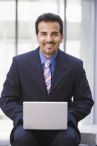 Businessman sitting outdoors by building with a laptop smiling (high key/selective focus)