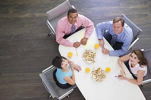Four businesspeople at boardroom table with sandwiches smiling