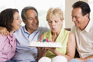 Two couples in living room smiling with woman blowing out candles on cake