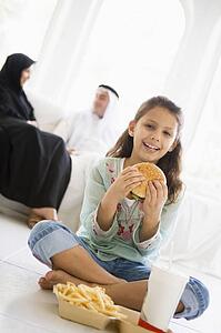 Young girl with fast food in living room smiling with parents in background (high key/selective focus)