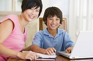 Woman helping young boy with laptop do homework in dining room smiling