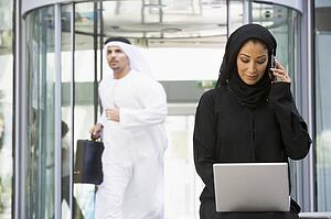 Businesswoman sitting outdoors by building with laptop using cellular phone with businessman running in background (selective focus)
