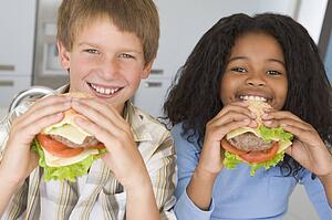 Two young children in kitchen eating cheeseburgers smiling