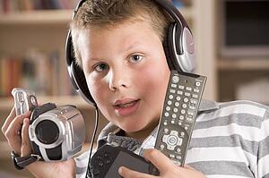 Young boy wearing headphones in bedroom holding many electronic devices