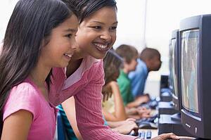 Teacher helping student at computer terminal with students in background (depth of field/high key)