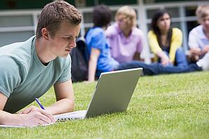 Student outdoors on lawn using laptop with other students in background (selective focus)