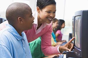 Teacher helping student at computer terminal with students in background (depth of field/high key)