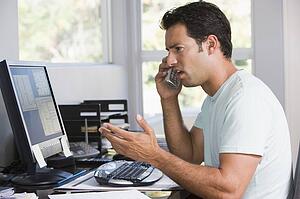 Man in home office on telephone using computer and frowning