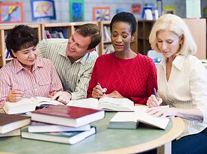 Three women sitting in library with books and notepads while a man leans over them (selective focus)