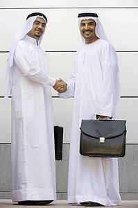 Two businessmen standing outdoors with briefcases shaking hands and smiling