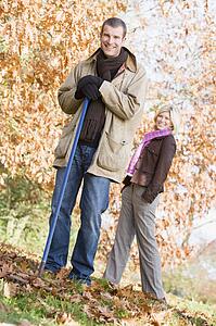 Man outdoors raking leaves and woman in background smiling (selective focus)