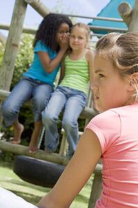 Two young girl friends at a playground whispering about other girl in foreground