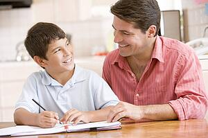 Man helping young boy in kitchen doing homework and smiling