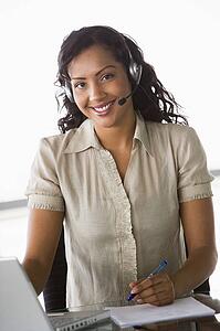 Businesswoman wearing headset in office by laptop smiling (high key/selective focus)
