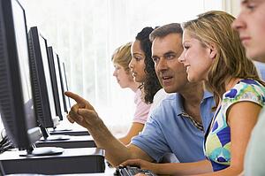 Four students sitting at computer terminals with teacher helping one of them (depth of field/high key)