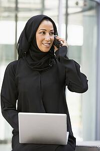 Businesswoman sitting outdoors by building with laptop using cellular phone smiling (selective focus)