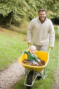 Man walking on path outdoors pushing baby in wheelbarrow and smiling (selective focus)