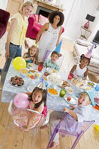 Young children at party with mothers sitting at table with food smiling