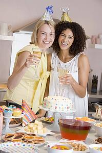 Two women at party holding drinks standing by food table smiling