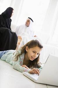 Young girl in living room using laptop and smiling with parents in background (high key/selective focus)