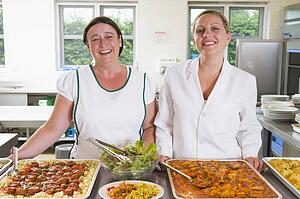 Two lunch ladies standing behind full lunch service station