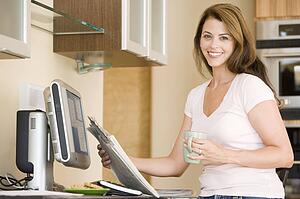 Woman in kitchen at computer with newspaper and coffee smiling