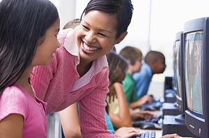 Teacher helping student at computer terminal with students in background (depth of field/high key)
