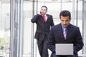Businessman outdoors in front of building using laptop with businessman in background on cellular phone (high key/selective focus)