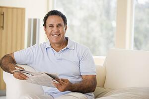 Man in living room reading newspaper smiling