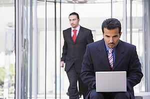 Businessman outdoors in front of building using laptop with businessman in background (high key/selective focus)