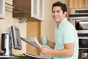 Man in kitchen with computer holding newspaper and coffee smiling