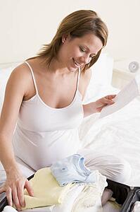 Pregnant woman packing baby clothing in suitcase holding list smiling