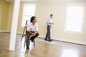 Man sitting on ladder in empty space with another man holding paper