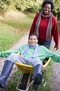 Mother outdoors pushing son in wheelbarrow and smiling (selective focus)