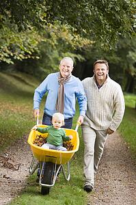 Two men walking on path outdoors pushing baby in wheelbarrow and smiling (selective focus)