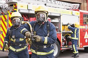 Two firefighters with hose and axe walking away from fire engine and another firefighter in background (selective focus)