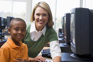Teacher sitting with student at computer terminal with students in background (selective focus/high key)